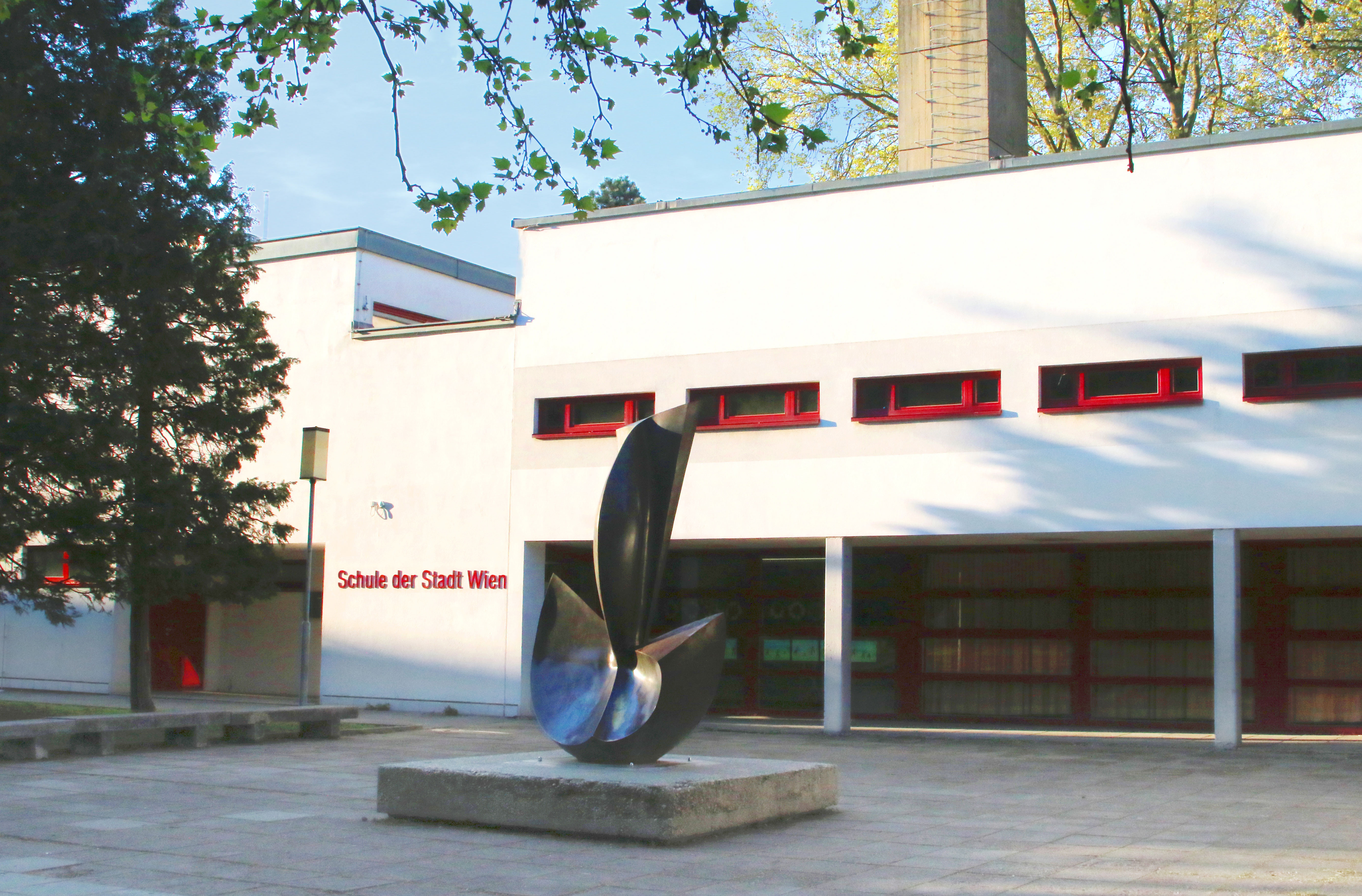 Weiß gefasste Eingangsfassade der Volksschule von Gustav Peichl mit roten Fensterrahmen und abstrakter Plastik von Wander Bertoni